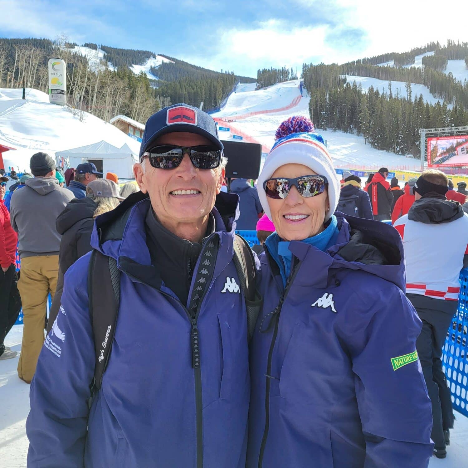 Debby and Harry Jasper smile while wearing beanies, puffer coats and sunglasses on a winter day with the ski racing course behind them